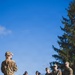 A military briefing or instruction session taking place outdoors on a grassy field under a clear blue sky. One individual in OCP (Operational Camouflage Pattern) uniform faces a group of approximately 8-10 military personnel, some in OCP uniforms and others in dark flight suits. The setting and attire suggest a U.S. Air Force training or operational exercise.