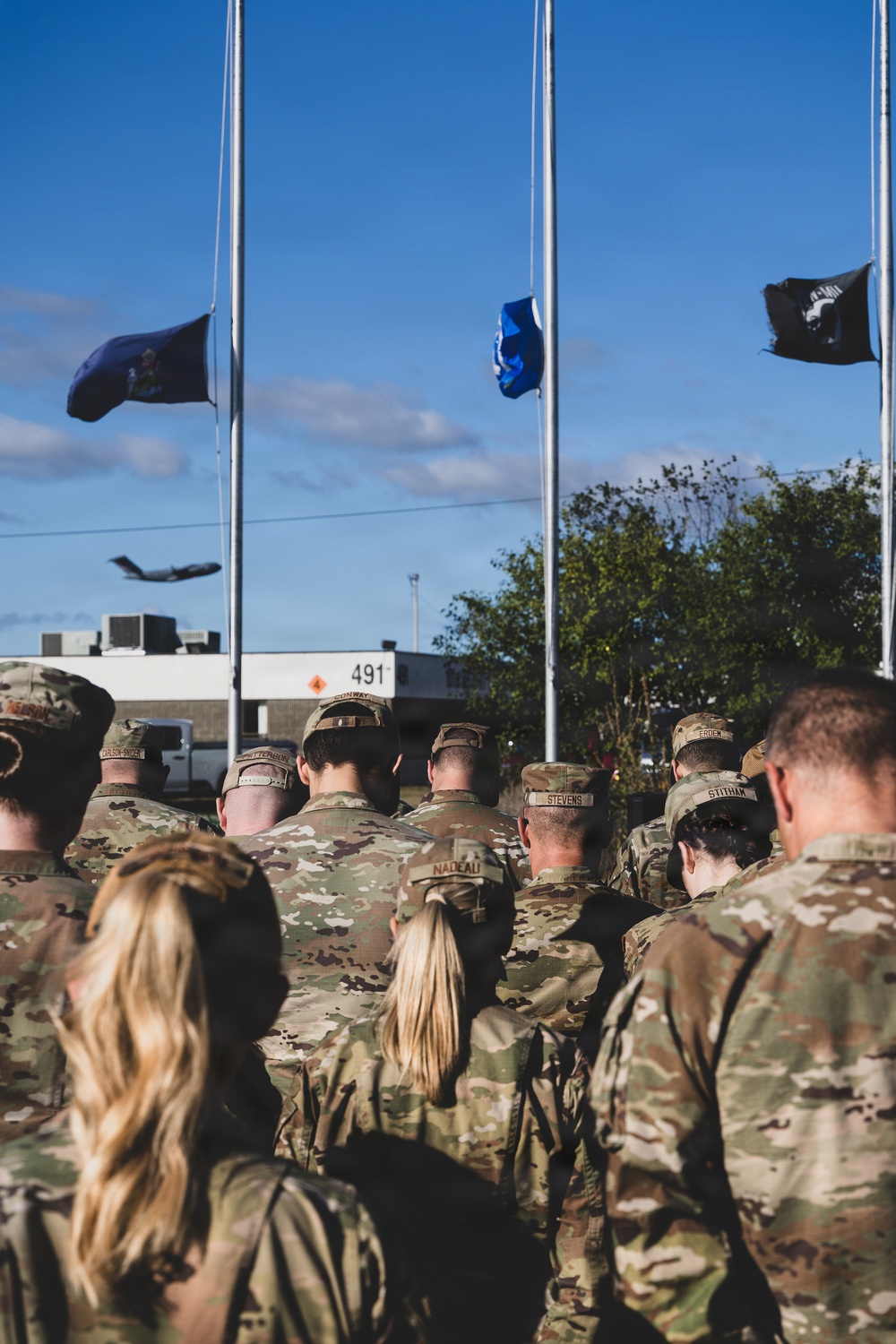 A group of military personnel in OCP (Operational Camouflage Pattern) uniforms are assembled outdoors facing multiple flagpoles during what appears to be a formal ceremony. Three flags are visible at half-staff, including what appears to be a state flag, a blue military branch flag, and a black POW/MIA flag. A military aircraft is visible in the background sky, suggesting proximity to an air base.