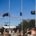 A group of military personnel in OCP (Operational Camouflage Pattern) uniforms are assembled outdoors facing multiple flagpoles during what appears to be a formal ceremony. Three flags are visible at half-staff, including what appears to be a state flag, a blue military branch flag, and a black POW/MIA flag. A military aircraft is visible in the background sky, suggesting proximity to an air base.
