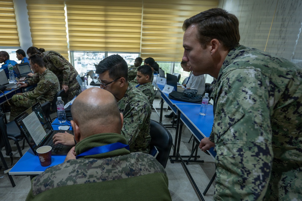Military personnel from multiple nations are gathered in a classroom or conference room setting, working on laptops at blue-covered tables. The scene appears to be a joint military training exercise, workshop, or planning session with participants wearing different camouflage uniforms. A US Navy sailor in NWU Type III digital camouflage is prominently visible observing or instructing others.