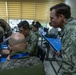 Military personnel from multiple nations are gathered in a classroom or conference room setting, working on laptops at blue-covered tables. The scene appears to be a joint military training exercise, workshop, or planning session with participants wearing different camouflage uniforms. A US Navy sailor in NWU Type III digital camouflage is prominently visible observing or instructing others.