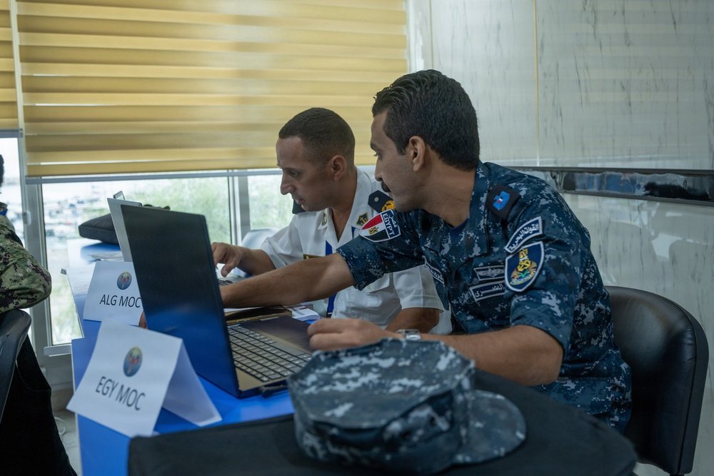 Two military personnel in uniform are collaborating at a laptop computer during what appears to be a multinational naval exercise or coordination meeting. Desk placards identify their stations as 'EGY MOC' (Egypt Maritime Operations Center) and 'ALG MOC' (Algeria Maritime Operations Center). The setting appears to be a conference or operations room with other uniformed personnel visible in the background.
