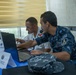 Two military personnel in uniform are collaborating at a laptop computer during what appears to be a multinational naval exercise or coordination meeting. Desk placards identify their stations as 'EGY MOC' (Egypt Maritime Operations Center) and 'ALG MOC' (Algeria Maritime Operations Center). The setting appears to be a conference or operations room with other uniformed personnel visible in the background.