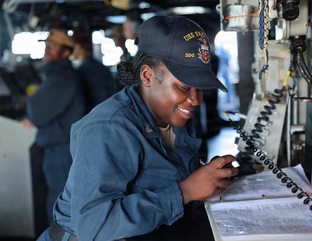 A U.S. Navy sailor is working at a station aboard a naval vessel, smiling while writing in or reviewing a binder/logbook. She is wearing a Navy working uniform (coveralls) and a cap bearing 'USS FAR...' and 'DDG' insignia, indicating she is assigned to a guided-missile destroyer. The setting appears to be the bridge or a command station of the ship.