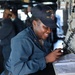 A U.S. Navy sailor is working at a station aboard a naval vessel, smiling while writing in or reviewing a binder/logbook. She is wearing a Navy working uniform (coveralls) and a cap bearing 'USS FAR...' and 'DDG' insignia, indicating she is assigned to a guided-missile destroyer. The setting appears to be the bridge or a command station of the ship.