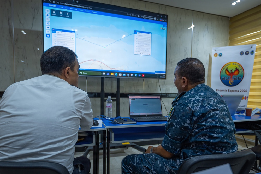 Two individuals — one in civilian attire and one in military camouflage uniform — are seated at a table reviewing a maritime tracking or navigation software displayed on a large screen. The setting appears to be a briefing or operations room associated with the 'Phoenix Express 2026' exercise, as indicated by a banner visible on the right. The screen displays what appears to be a vessel tracking or route-planning application (SPLUSS) with map overlays.