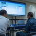 Two individuals — one in civilian attire and one in military camouflage uniform — are seated at a table reviewing a maritime tracking or navigation software displayed on a large screen. The setting appears to be a briefing or operations room associated with the 'Phoenix Express 2026' exercise, as indicated by a banner visible on the right. The screen displays what appears to be a vessel tracking or route-planning application (SPLUSS) with map overlays.