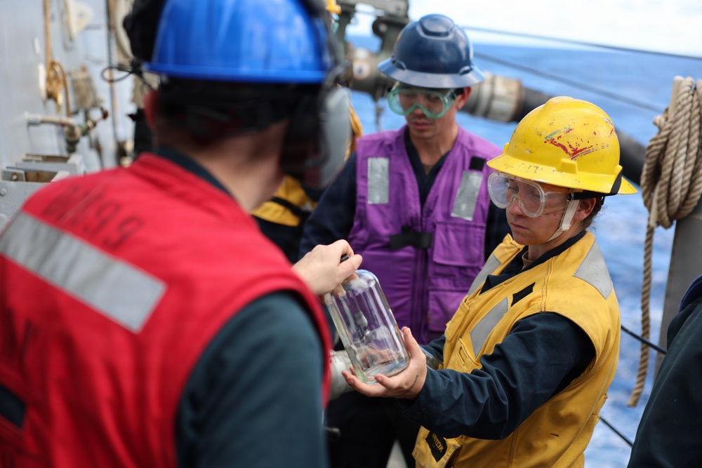 Three workers in personal protective equipment are gathered on what appears to be the deck of a naval or maritime vessel. A worker in a yellow hard hat and safety goggles is holding a clear glass jar or container, appearing to examine or present it to a colleague in a red vest. The ocean is visible in the background, confirming an at-sea location.