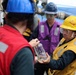 Three workers in personal protective equipment are gathered on what appears to be the deck of a naval or maritime vessel. A worker in a yellow hard hat and safety goggles is holding a clear glass jar or container, appearing to examine or present it to a colleague in a red vest. The ocean is visible in the background, confirming an at-sea location.