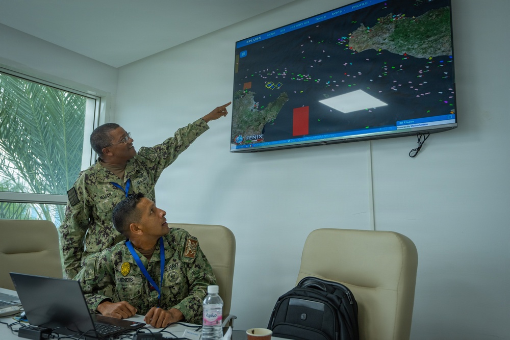 Two U.S. Navy personnel in digital camouflage uniforms are reviewing a maritime tracking display system called 'FENIX' mounted on a wall. One standing officer, identified by nametape as 'NELSON,' is pointing at the screen while a seated officer identified as 'MENDIS' observes. The screen displays what appears to be a maritime domain awareness or vessel tracking system showing a coastal region with multiple colored vessel markers.