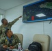 Two U.S. Navy personnel in digital camouflage uniforms are reviewing a maritime tracking display system called 'FENIX' mounted on a wall. One standing officer, identified by nametape as 'NELSON,' is pointing at the screen while a seated officer identified as 'MENDIS' observes. The screen displays what appears to be a maritime domain awareness or vessel tracking system showing a coastal region with multiple colored vessel markers.