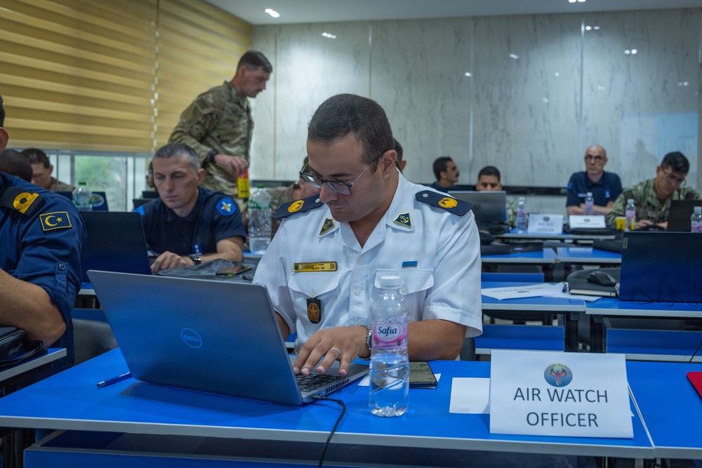 A military training or briefing session is taking place in a conference room with personnel from multiple nations. The foreground shows an officer designated as 'Air Watch Officer' working on a Dell laptop. Multiple uniformed personnel from different military branches are seated at blue tables with laptops and water bottles.