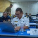 A military training or briefing session is taking place in a conference room with personnel from multiple nations. The foreground shows an officer designated as 'Air Watch Officer' working on a Dell laptop. Multiple uniformed personnel from different military branches are seated at blue tables with laptops and water bottles.