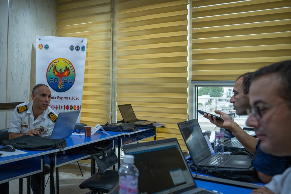 A military briefing or planning session is taking place in a conference room, associated with the 'Phoenix Express 2026' multinational exercise. A uniformed military officer sits at a table with a laptop, facing two civilians or other participants who are also working on laptops and a tablet. The banner in the background displays the Phoenix Express 2026 logo along with multiple national flags.