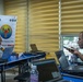 A military briefing or planning session is taking place in a conference room, associated with the 'Phoenix Express 2026' multinational exercise. A uniformed military officer sits at a table with a laptop, facing two civilians or other participants who are also working on laptops and a tablet. The banner in the background displays the Phoenix Express 2026 logo along with multiple national flags.