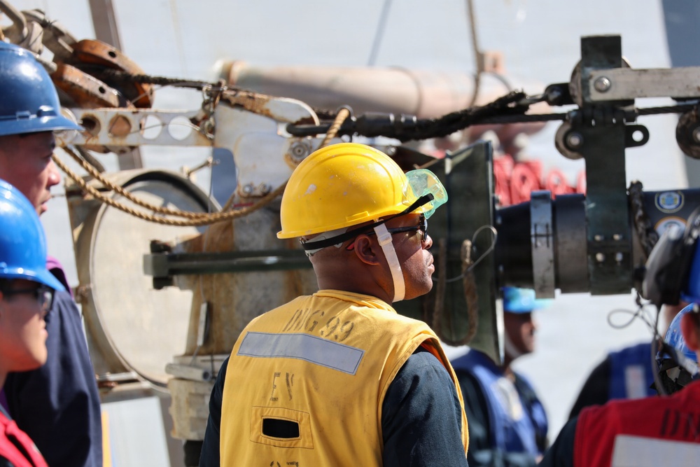 The image shows a group of workers on what appears to be a ship or industrial maritime vessel. The central figure is wearing a yellow hard hat, safety goggles, and a yellow high-visibility vest with partial text reading 'DDG 99' and 'EV'. Multiple workers with different colored hard hats are visible in the background amid heavy industrial equipment.