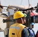 The image shows a group of workers on what appears to be a ship or industrial maritime vessel. The central figure is wearing a yellow hard hat, safety goggles, and a yellow high-visibility vest with partial text reading 'DDG 99' and 'EV'. Multiple workers with different colored hard hats are visible in the background amid heavy industrial equipment.