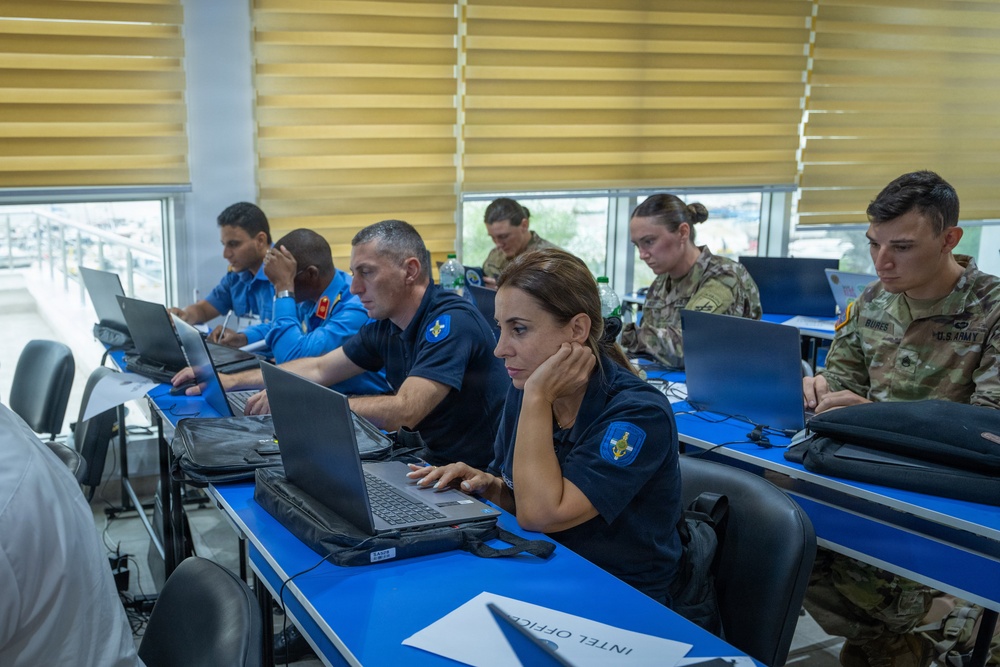 A mixed group of military and law enforcement personnel are seated at blue tables working on laptops in what appears to be a training or operations room. Participants include individuals in U.S. Army combat uniforms and others wearing blue uniforms with a star-badge shoulder patch. A placard reading 'INTEL OFFICE' is visible on one of the tables.