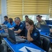 A mixed group of military and law enforcement personnel are seated at blue tables working on laptops in what appears to be a training or operations room. Participants include individuals in U.S. Army combat uniforms and others wearing blue uniforms with a star-badge shoulder patch. A placard reading 'INTEL OFFICE' is visible on one of the tables.