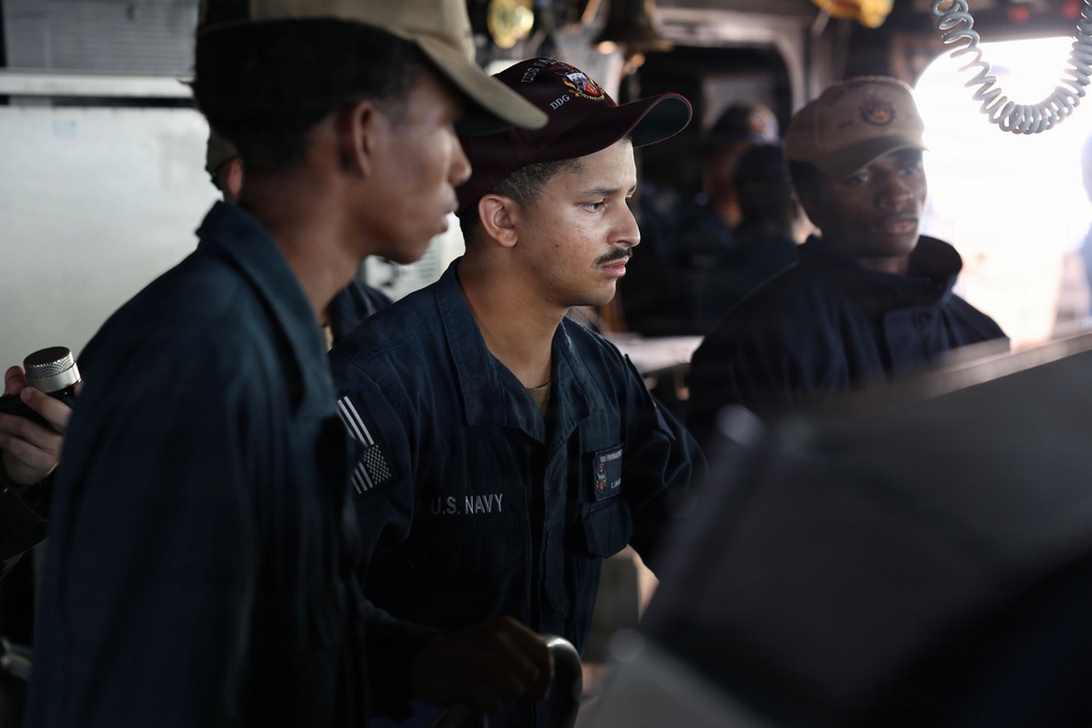 The image shows U.S. Navy personnel aboard what appears to be a naval vessel's bridge or combat information center. At least three sailors in dark navy working uniforms and caps are visible, with one centrally positioned sailor wearing a cap marked 'DDG' (destroyer designation). The personnel appear to be monitoring equipment or standing watch at their stations.