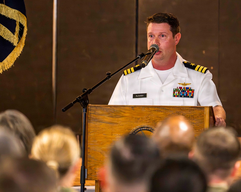 A U.S. Navy officer identified by nameplate as 'HLOUSEK' is speaking at a podium during a formal military ceremony. He is wearing a white Navy service uniform with gold shoulder boards indicating the rank of Captain (O-6), along with aviator wings and multiple service ribbons. The podium bears the insignia of 'Camp Lemonnier,' and an audience is visible in the foreground.