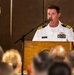 A U.S. Navy officer identified by nameplate as 'HLOUSEK' is speaking at a podium during a formal military ceremony. He is wearing a white Navy service uniform with gold shoulder boards indicating the rank of Captain (O-6), along with aviator wings and multiple service ribbons. The podium bears the insignia of 'Camp Lemonnier,' and an audience is visible in the foreground.