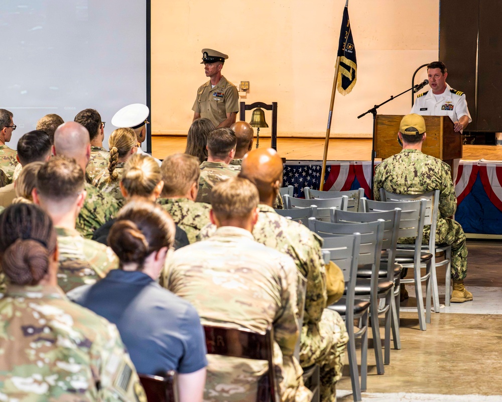 A military ceremony or briefing is taking place in an indoor venue, with a naval officer speaking at a podium to an audience of seated military personnel in various uniforms. The setting includes patriotic decorations, a ceremonial bell, and a military unit flag on stage. The audience consists of personnel from multiple branches, primarily wearing Army combat uniforms and Navy working uniforms.