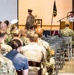 A military ceremony or briefing is taking place in an indoor venue, with a naval officer speaking at a podium to an audience of seated military personnel in various uniforms. The setting includes patriotic decorations, a ceremonial bell, and a military unit flag on stage. The audience consists of personnel from multiple branches, primarily wearing Army combat uniforms and Navy working uniforms.