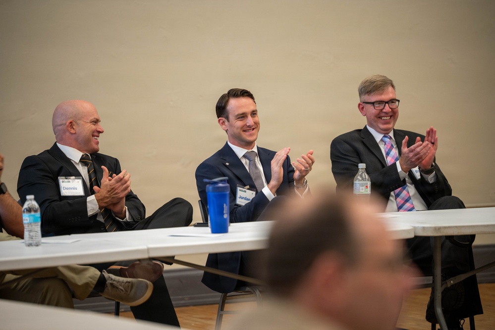 Three men in business attire are seated at a white folding table, smiling and applauding during what appears to be a professional panel or conference event. Name badges are visible on two of the men, with readable names 'Dennis' and 'Jordan'. The atmosphere is positive and celebratory.