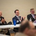 Three men in business attire are seated at a white folding table, smiling and applauding during what appears to be a professional panel or conference event. Name badges are visible on two of the men, with readable names 'Dennis' and 'Jordan'. The atmosphere is positive and celebratory.