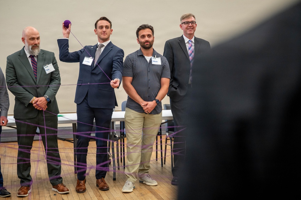 Four men wearing name tags stand in a line during what appears to be a team-building or networking activity involving a ball of purple yarn. The man second from left, identified as 'Jordan,' is holding the purple yarn ball aloft while strings of purple yarn crisscross in the foreground, suggesting a web-building exercise. The setting appears to be a gymnasium or large meeting hall with folding tables visible in the background.