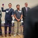 Four men wearing name tags stand in a line during what appears to be a team-building or networking activity involving a ball of purple yarn. The man second from left, identified as 'Jordan,' is holding the purple yarn ball aloft while strings of purple yarn crisscross in the foreground, suggesting a web-building exercise. The setting appears to be a gymnasium or large meeting hall with folding tables visible in the background.