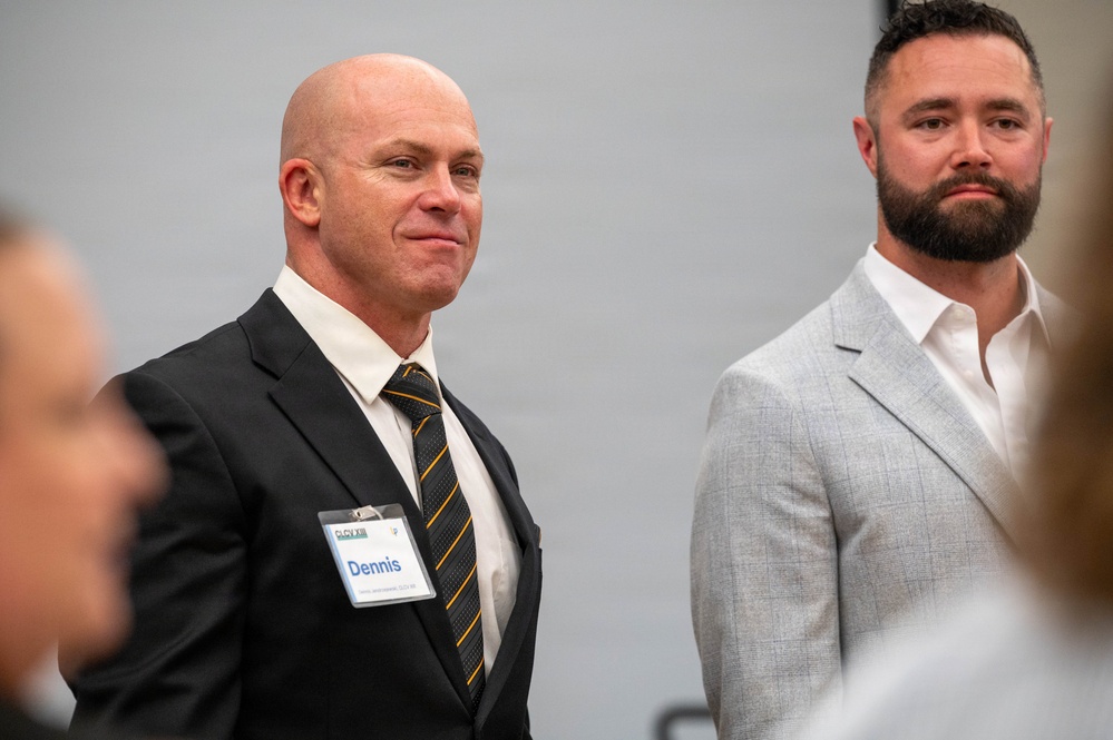 Two men in business attire are standing at what appears to be a professional event or conference. The man on the left is bald, wearing a dark suit with a striped tie, and has a name badge reading 'Dennis'. The man on the right has a full beard and is wearing a light gray suit with an open-collar white shirt.