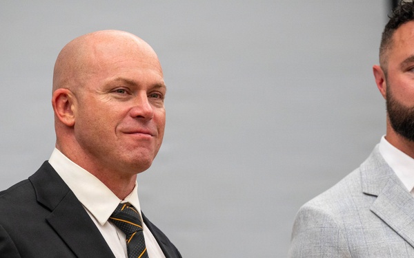 Two men in business attire are standing at what appears to be a professional event or conference. The man on the left is bald, wearing a dark suit with a striped tie, and has a name badge reading 'Dennis'. The man on the right has a full beard and is wearing a light gray suit with an open-collar white shirt.
