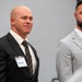 Two men in business attire are standing at what appears to be a professional event or conference. The man on the left is bald, wearing a dark suit with a striped tie, and has a name badge reading 'Dennis'. The man on the right has a full beard and is wearing a light gray suit with an open-collar white shirt.