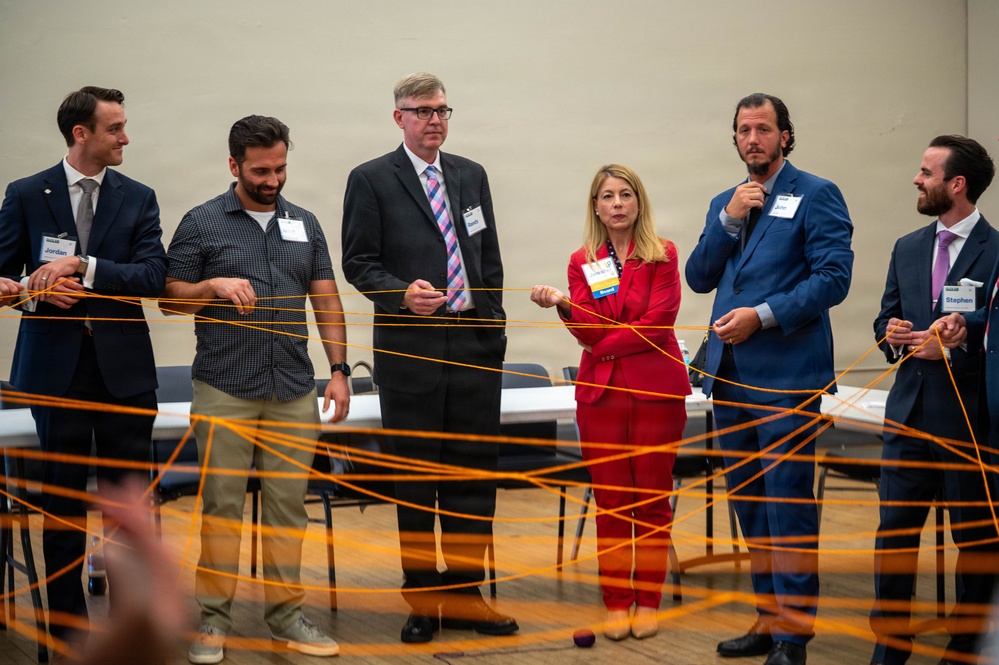 Six professionals wearing name badges are participating in a group activity or team-building exercise involving an orange string or yarn network. They are standing in a semi-circle in what appears to be a conference or event hall, each holding portions of the interconnected orange string. The activity appears to simulate a web of connections, commonly used in networking or leadership workshops.