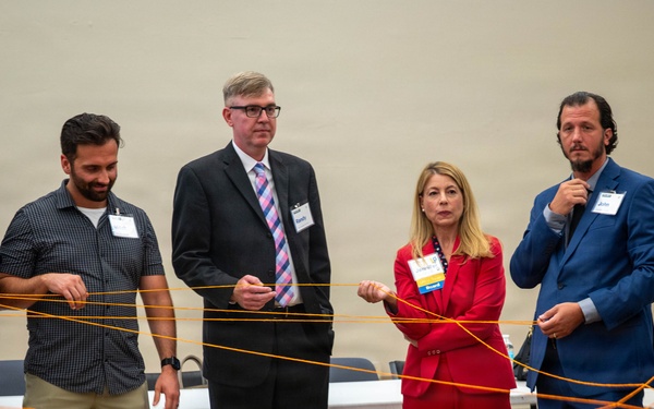 Six professionals wearing name badges are participating in a group activity or team-building exercise involving an orange string or yarn network. They are standing in a semi-circle in what appears to be a conference or event hall, each holding portions of the interconnected orange string. The activity appears to simulate a web of connections, commonly used in networking or leadership workshops.