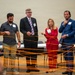 Six professionals wearing name badges are participating in a group activity or team-building exercise involving an orange string or yarn network. They are standing in a semi-circle in what appears to be a conference or event hall, each holding portions of the interconnected orange string. The activity appears to simulate a web of connections, commonly used in networking or leadership workshops.
