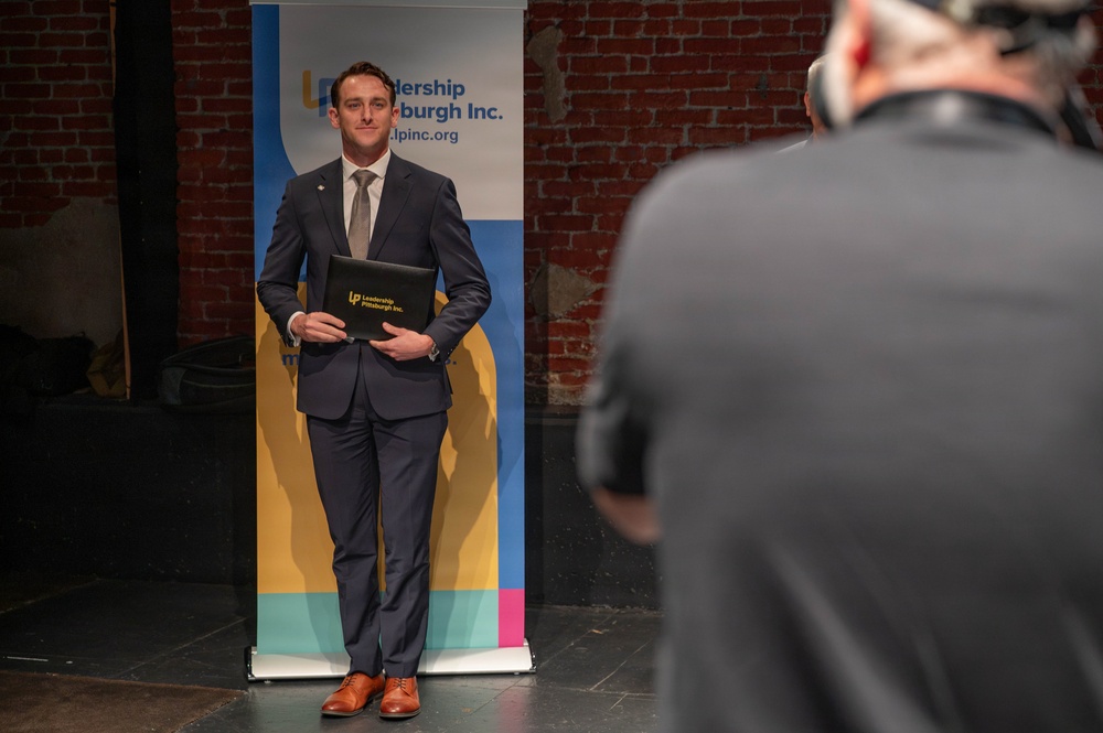 A man in a navy blue suit is posing for a photo in front of a 'Leadership Pittsburgh Inc.' branded banner. He is holding a black portfolio/folder embossed with the Leadership Pittsburgh Inc. logo. A photographer is visible in the foreground, partially out of focus, capturing the moment.