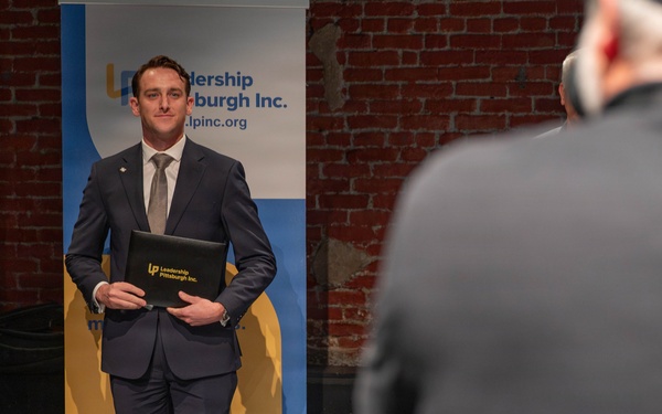 A man in a navy blue suit is posing for a photo in front of a 'Leadership Pittsburgh Inc.' branded banner. He is holding a black portfolio/folder embossed with the Leadership Pittsburgh Inc. logo. A photographer is visible in the foreground, partially out of focus, capturing the moment.