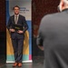 A man in a navy blue suit is posing for a photo in front of a 'Leadership Pittsburgh Inc.' branded banner. He is holding a black portfolio/folder embossed with the Leadership Pittsburgh Inc. logo. A photographer is visible in the foreground, partially out of focus, capturing the moment.