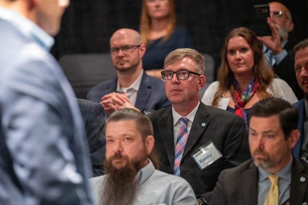 A group of attendees seated at what appears to be a professional conference or formal meeting. The central figure is a man wearing a black suit with a plaid tie and a name badge reading 'Randy,' attentively watching a presenter or speaker out of frame to the left. The audience appears engaged, with multiple attendees in business attire visible in the background.