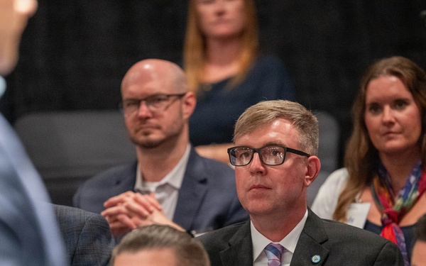A group of attendees seated at what appears to be a professional conference or formal meeting. The central figure is a man wearing a black suit with a plaid tie and a name badge reading 'Randy,' attentively watching a presenter or speaker out of frame to the left. The audience appears engaged, with multiple attendees in business attire visible in the background.