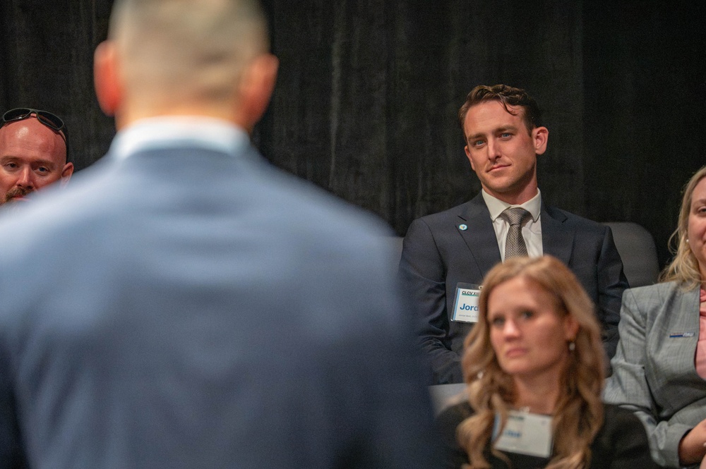 A formal event or conference setting where attendees are seated and listening to a speaker whose back is visible in the foreground. A young man in a dark suit with a name badge partially reading 'Jord...' is prominently visible, attentively watching the speaker. The dark backdrop and professional attire suggest a business or organizational gathering.