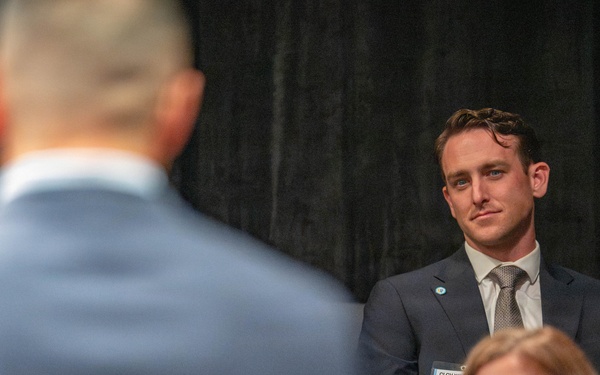 A formal event or conference setting where attendees are seated and listening to a speaker whose back is visible in the foreground. A young man in a dark suit with a name badge partially reading 'Jord...' is prominently visible, attentively watching the speaker. The dark backdrop and professional attire suggest a business or organizational gathering.