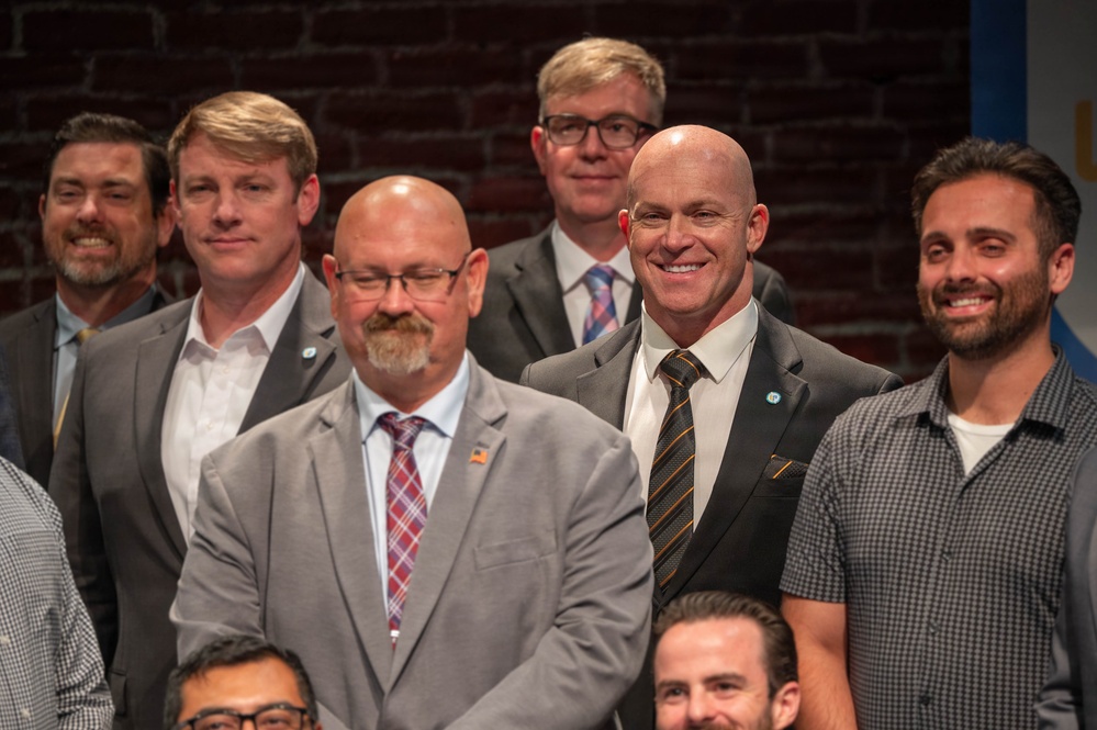 A group of men are posing together for a formal group photo, likely at a professional or civic event. The setting features an exposed brick wall background, suggesting an urban venue. The individuals are dressed in a mix of business formal and business casual attire.