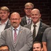 A group of men are posing together for a formal group photo, likely at a professional or civic event. The setting features an exposed brick wall background, suggesting an urban venue. The individuals are dressed in a mix of business formal and business casual attire.