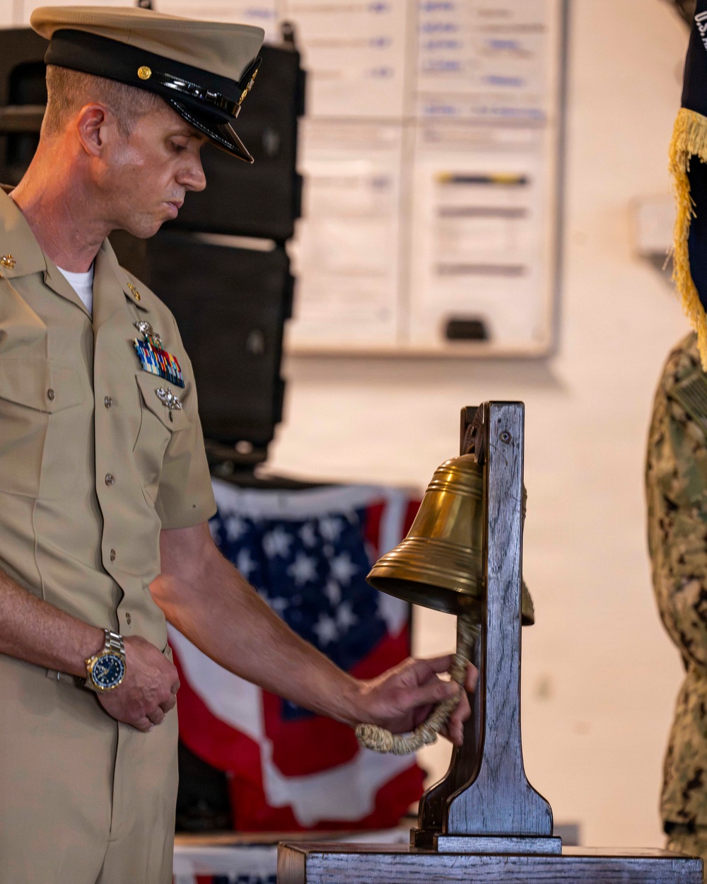 A U.S. Navy officer in service khaki uniform is ringing a brass ship's bell mounted on a wooden stand during what appears to be a formal military ceremony. The setting includes an American flag in the background and another service member in camouflage utilities visible at the right edge. The occasion appears to be a change of command, retirement, or similar naval ceremonial event.