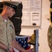 A U.S. Navy officer in service khaki uniform is ringing a brass ship's bell mounted on a wooden stand during what appears to be a formal military ceremony. The setting includes an American flag in the background and another service member in camouflage utilities visible at the right edge. The occasion appears to be a change of command, retirement, or similar naval ceremonial event.