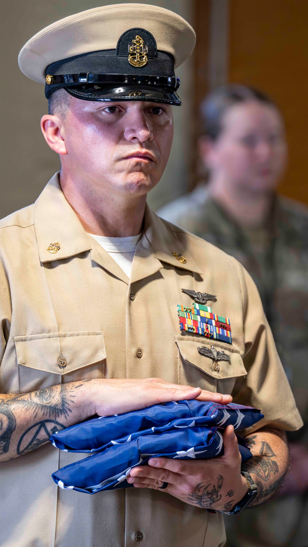 A U.S. Navy Chief Petty Officer in Service Khaki uniform stands at attention holding a folded American flag, suggesting a formal military ceremony such as a retirement or memorial service. The sailor displays multiple service ribbons, an aviation warfare device, and a surface warfare device on his uniform. A second uniformed individual is visible but out of focus in the background.